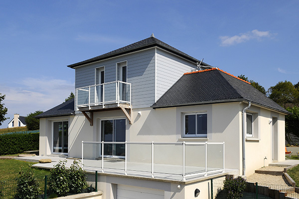 Maison avec extension moderne à deux étages, balcons en verre et toit en ardoise, sous un ciel bleu.