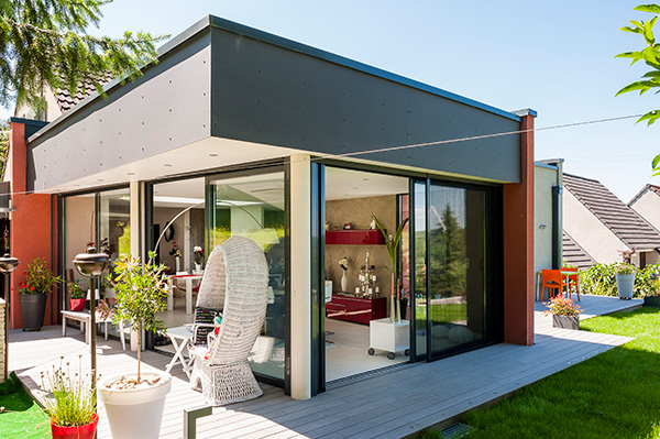 Extension moderne de maison avec grandes baies vitrées, terrasse en bois et mobilier extérieur dans un jardin verdoyant.