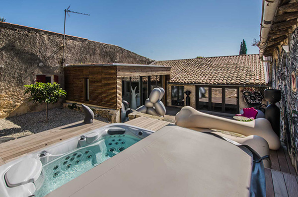 Maison moderne avec extension bois, grande terrasse et jacuzzi, entourée de murs en pierre sous un ciel bleu clair.