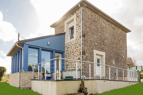 Maison en pierre rénovée avec extension moderne en bois bleu et terrasse, sous un ciel partiellement nuageux.