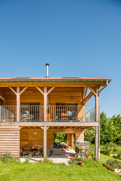 Maison en bois à deux étages avec balcon, située dans un jardin verdoyant sous un ciel bleu.