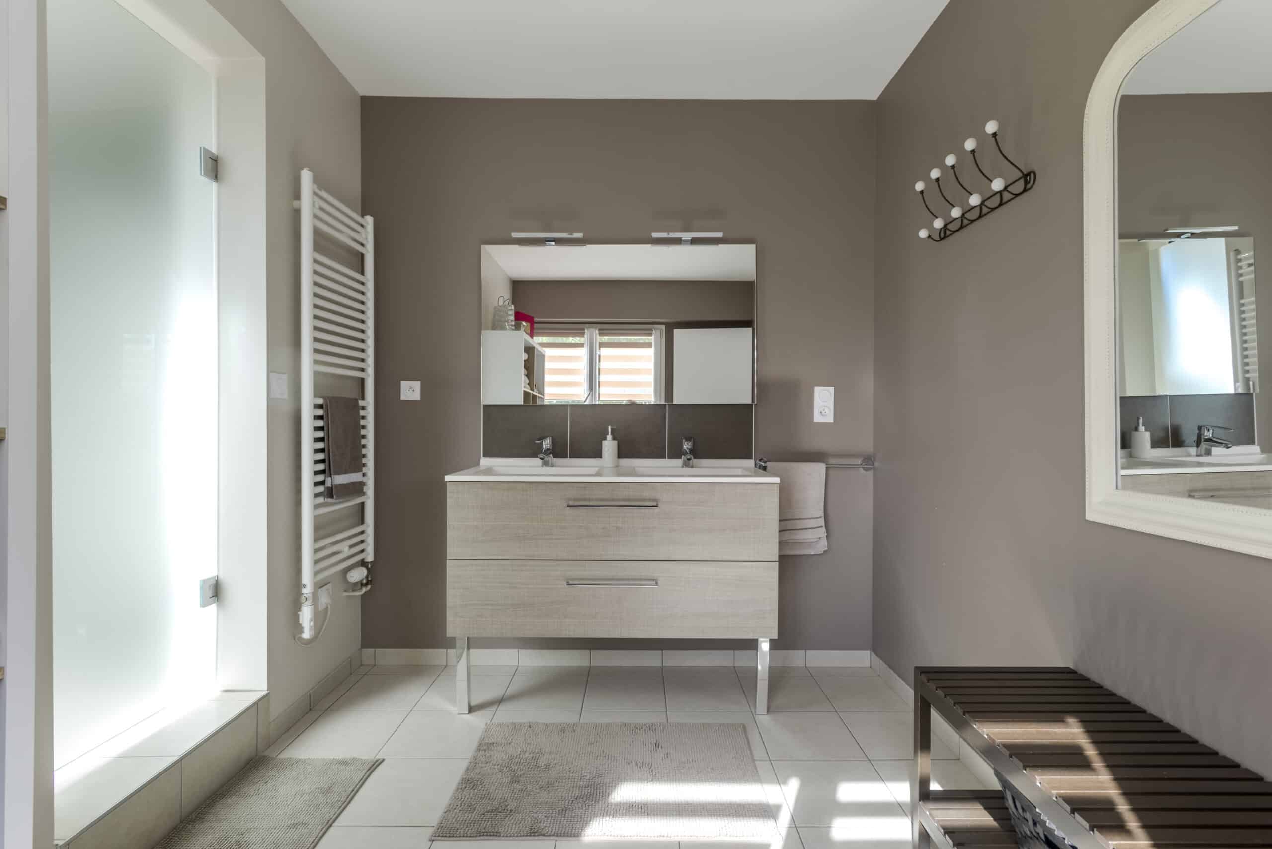 Salle de bain moderne avec double vasque, grand miroir, murs gris, et lumière naturelle provenant d